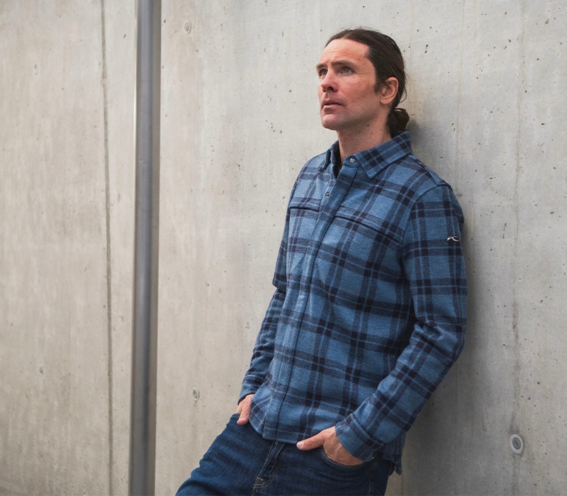 Man leaning against a concrete wall while wearing a blue plaid KJUS button-up shirt and jeans.